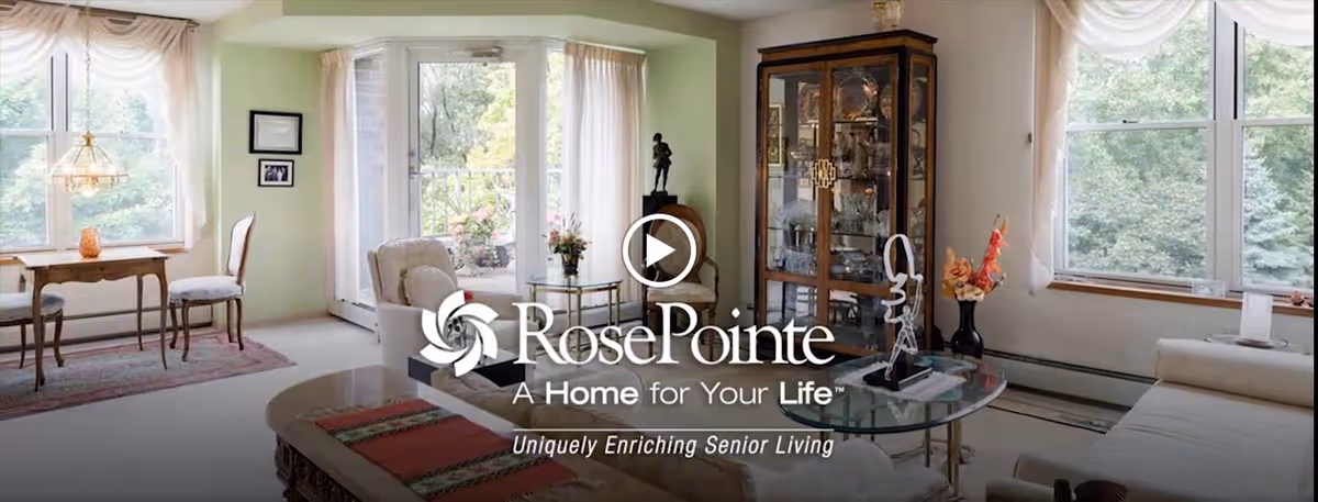 A bright and cozy living room area in a senior living facility with large windows letting in natural light. The room features a glass coffee table with decorative items, a wooden display cabinet filled with glassware, a small dining table with chairs near the windows, and comfortable seating including a sofa and armchairs. Light-colored curtains frame the windows and a door leading outside.