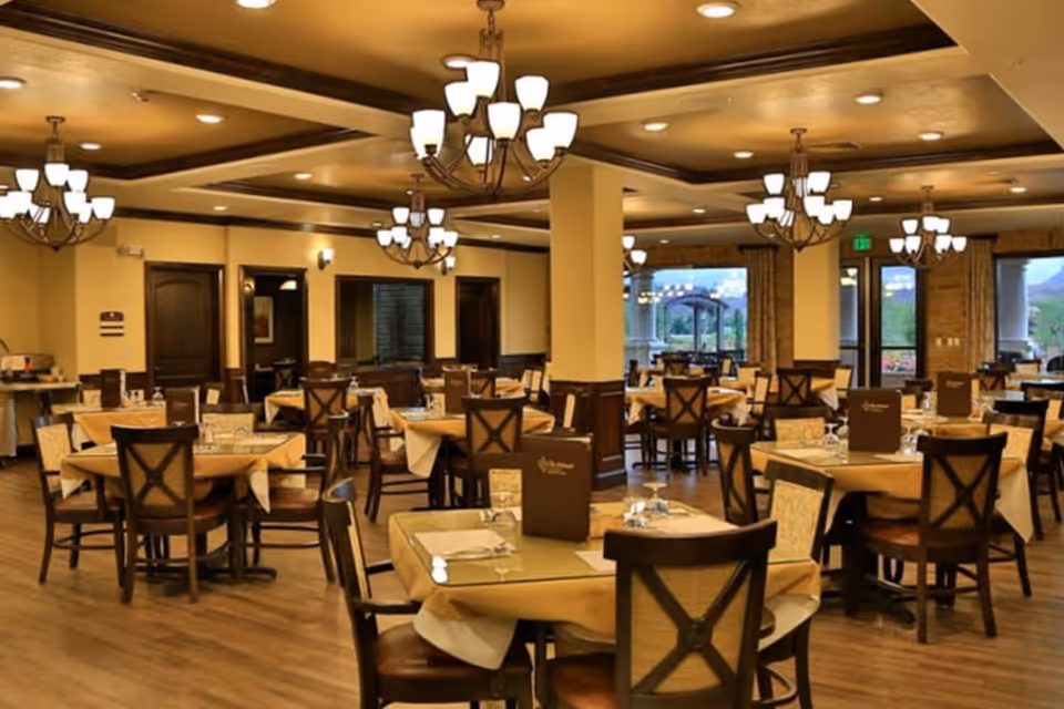 Spacious, warmly lit dining room with multiple round tables and wooden chairs set with place settings and chandeliers overhead.