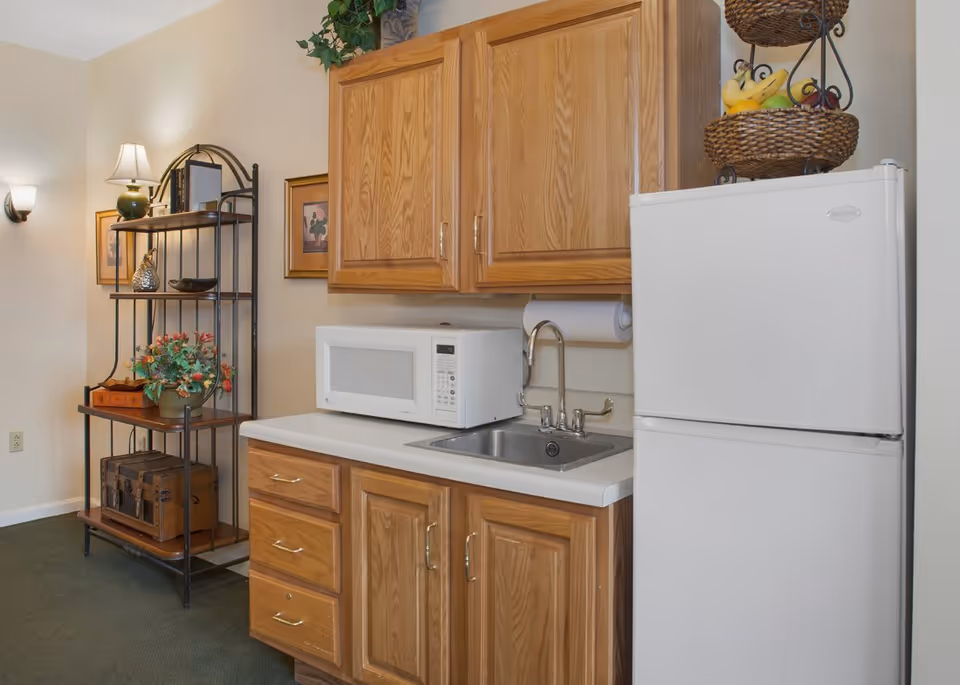 Small kitchenette with wooden cabinets, a microwave, sink, and white refrigerator beside a decorative shelving unit.
