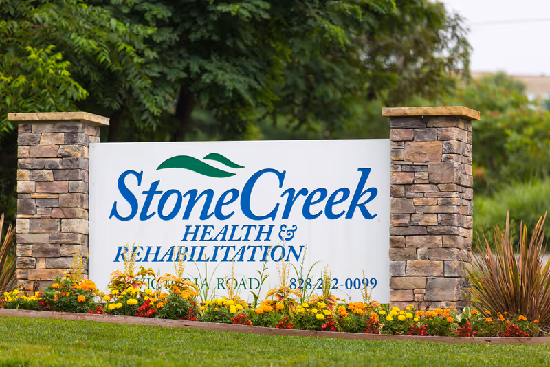 Stonecreek Health & Rehabilitation facility sign made of white board with blue and green text, flanked by two stone pillars and surrounded by colorful flowers and greenery.