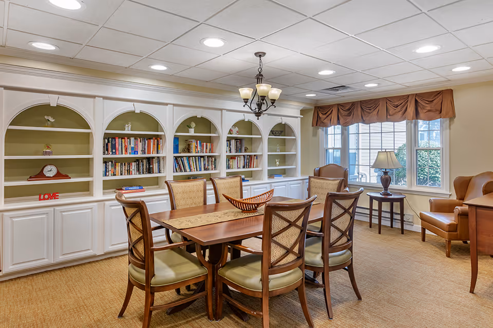 Bright communal dining and reading room with a wooden table and chairs, built-in white bookshelves, and windows with seating.