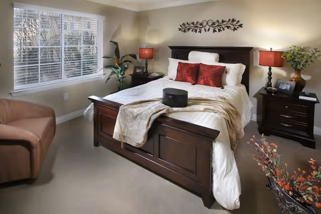 A cozy bedroom with a wooden bed frame, white bedding, and two red decorative pillows. There are two dark wooden nightstands on either side of the bed, each with a red lamp and decorative items. A beige armchair is positioned near a window with white blinds, and a potted plant is placed next to the bed. The room is softly lit and decorated with a wall ornament above the bed and a vase with flowers in the foreground.