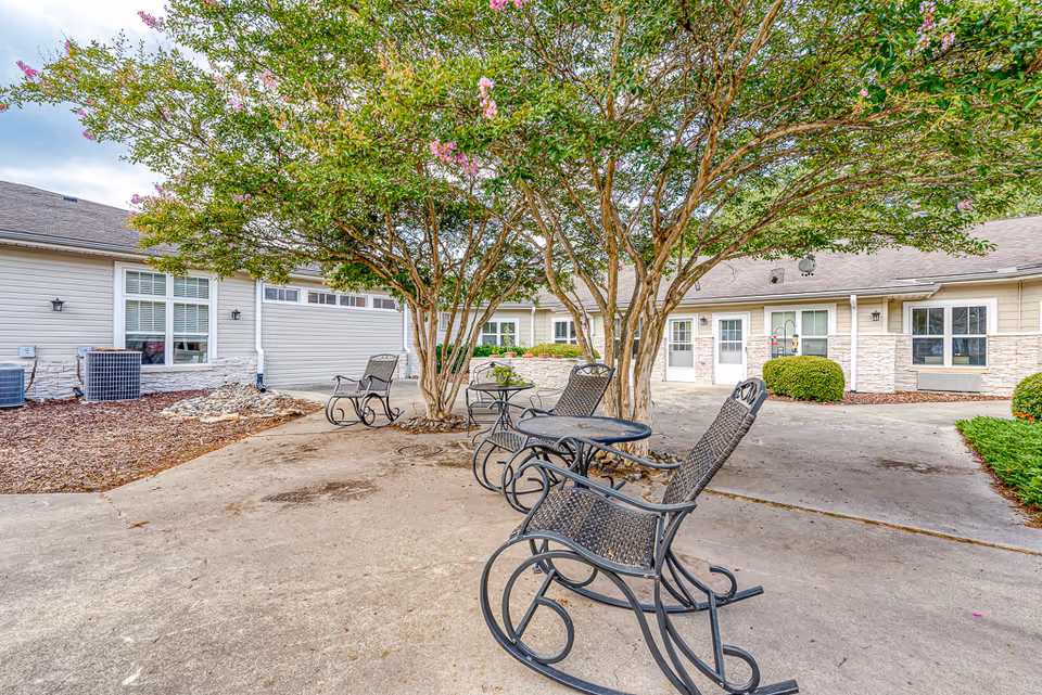 Outdoor courtyard with metal rocking chairs and tables under trees in front of a single-story senior living building.