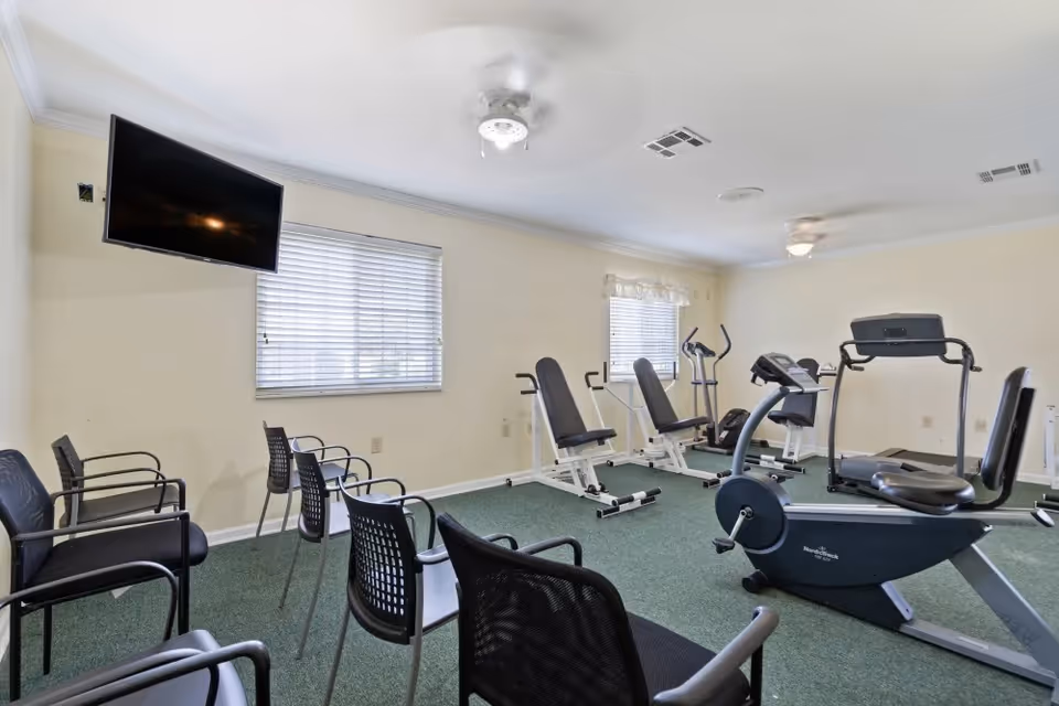 A fitness room with exercise equipment including stationary bikes, elliptical machines, and weight benches. Several black chairs are arranged along the walls. The room has beige walls, green carpet, two windows with blinds, and a wall-mounted flat-screen TV.
