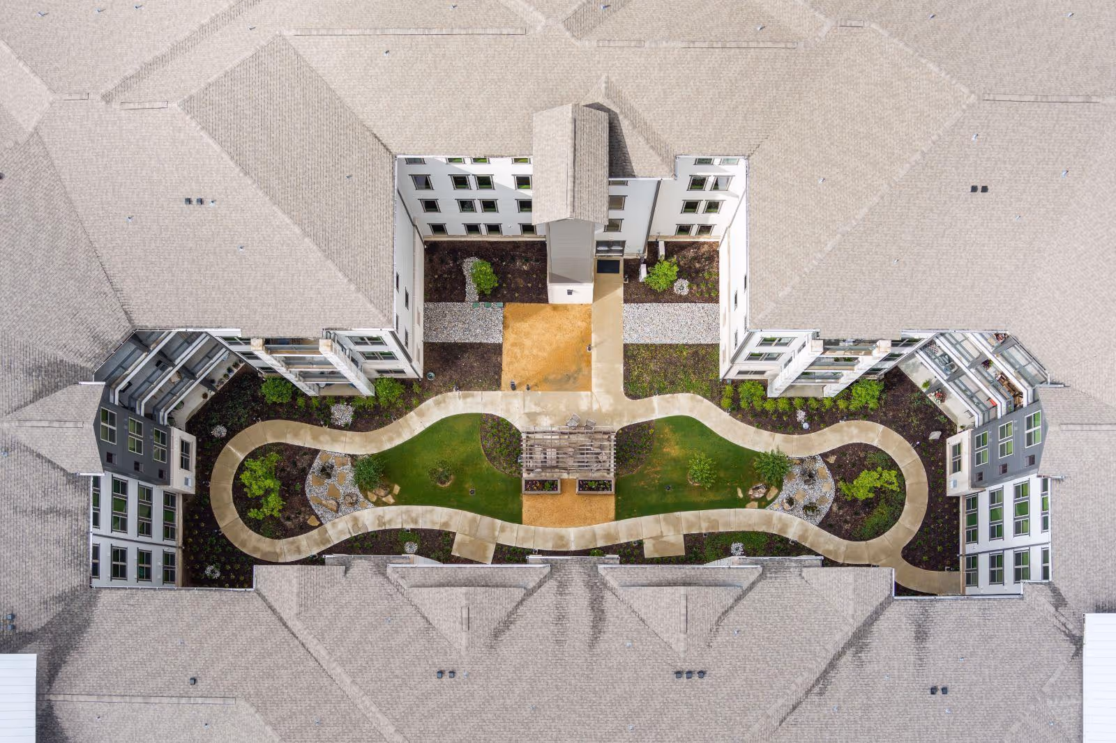 Aerial view of the courtyard of a senior living facility named Hidden Springs of McKinney, showing a landscaped garden with a winding concrete pathway, green grass, shrubs, and a wooden pergola in the center, surrounded by the building's roof and windows.