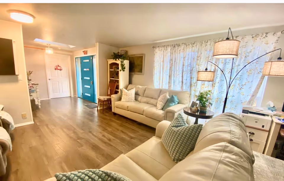 Bright, cozy living room with beige sofas, patterned curtains, a multi-arm floor lamp, and a teal front door visible at the entry.