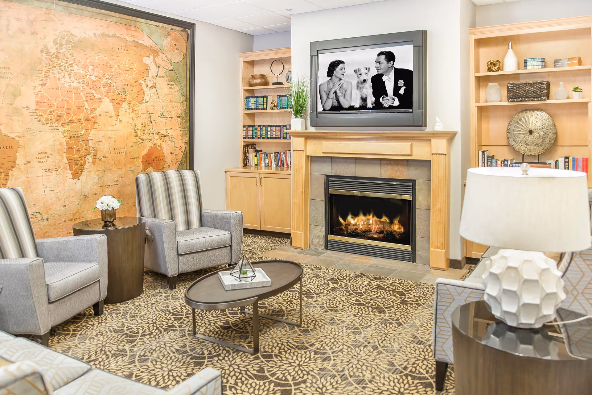A cozy living room with a patterned carpet, two striped armchairs, a small round table with a flower vase, a fireplace with a wooden mantel, and a TV above it displaying a black and white image of a man, woman, and dog. There are built-in wooden bookshelves on either side of the fireplace filled with books and decorative items. A large vintage-style world map hangs on the wall to the left.