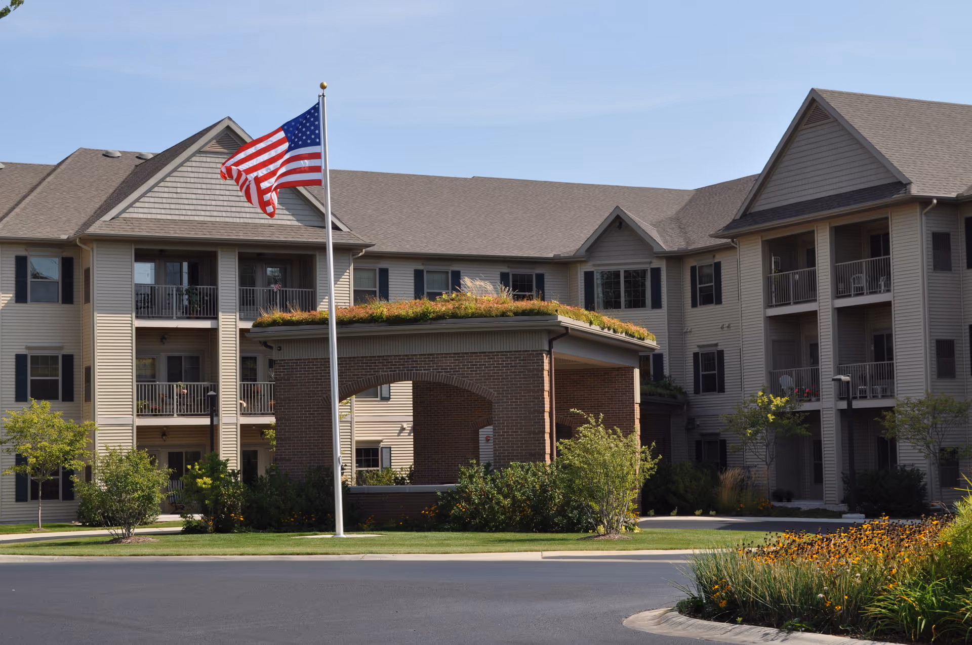 Exterior view of a multi-story senior living facility with beige siding and dark shutters. The building has balconies and a covered entrance with a green roof. An American flag is flying on a flagpole in front of the building, surrounded by landscaped greenery and flowers.
