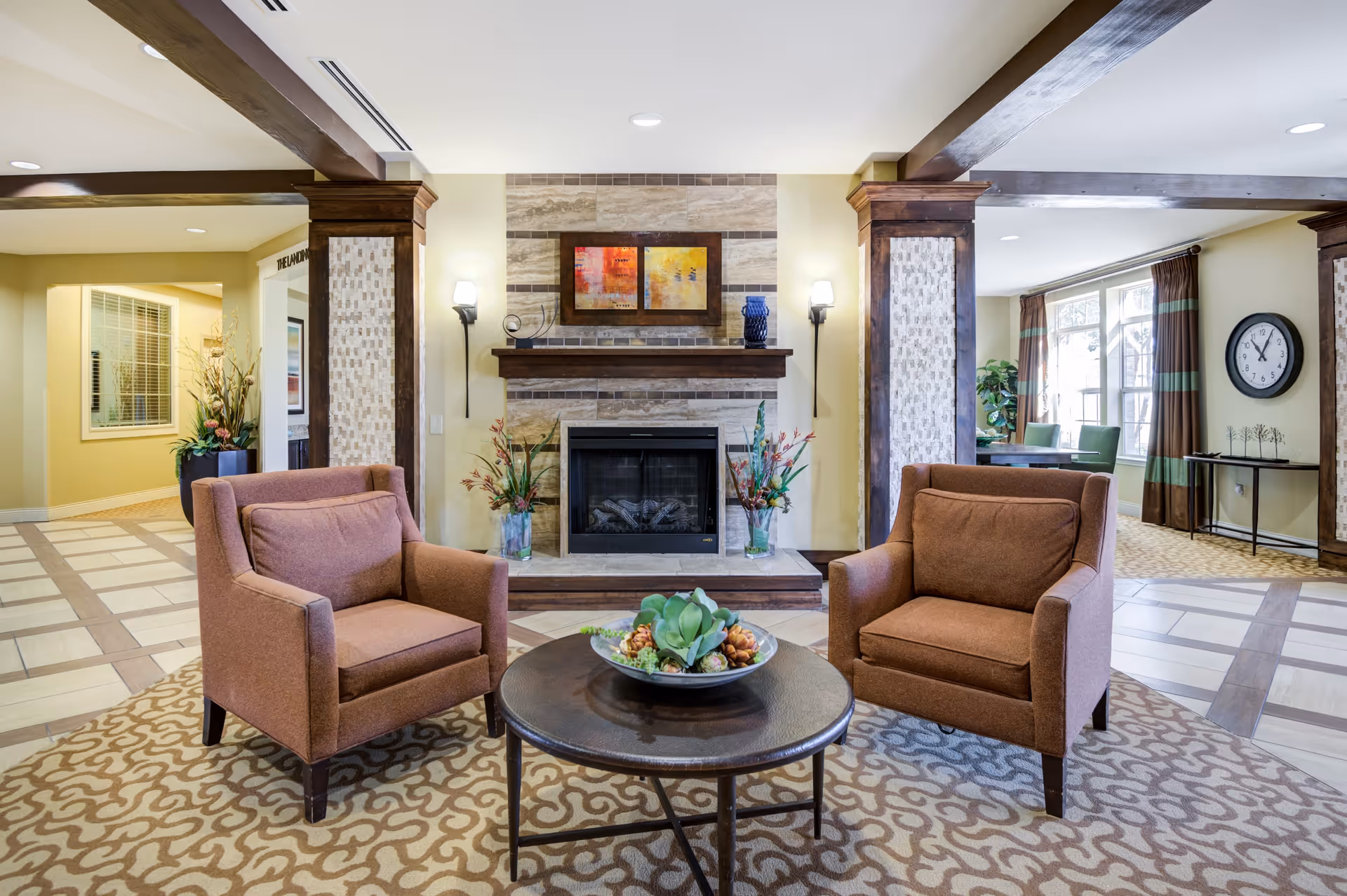 A cozy living room area with two brown armchairs facing a round wooden coffee table with a decorative centerpiece. Behind the chairs is a fireplace with a stone surround, flanked by two wall sconces and decorated with two vases of flowers and framed artwork above the mantel. The room features wooden beams, patterned carpet, and large windows with curtains letting in natural light.