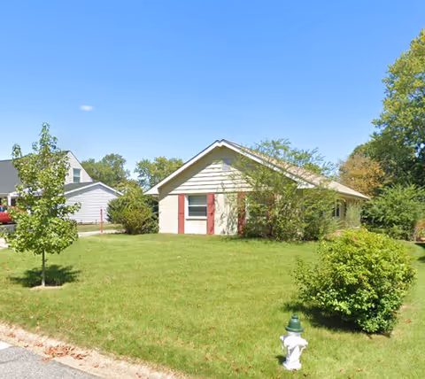 Single-story residential building with beige siding and red shutters, surrounded by green grass, bushes, and trees under a clear blue sky. A white fire hydrant is visible on the grass near the street curb.