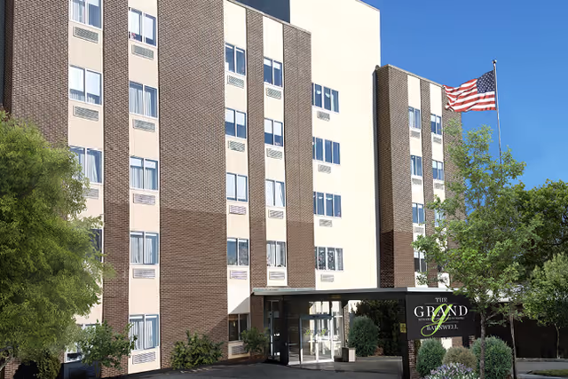 Front exterior of a multi-story brick-and-stucco senior living building with an entrance canopy, landscaping, and an American flag.