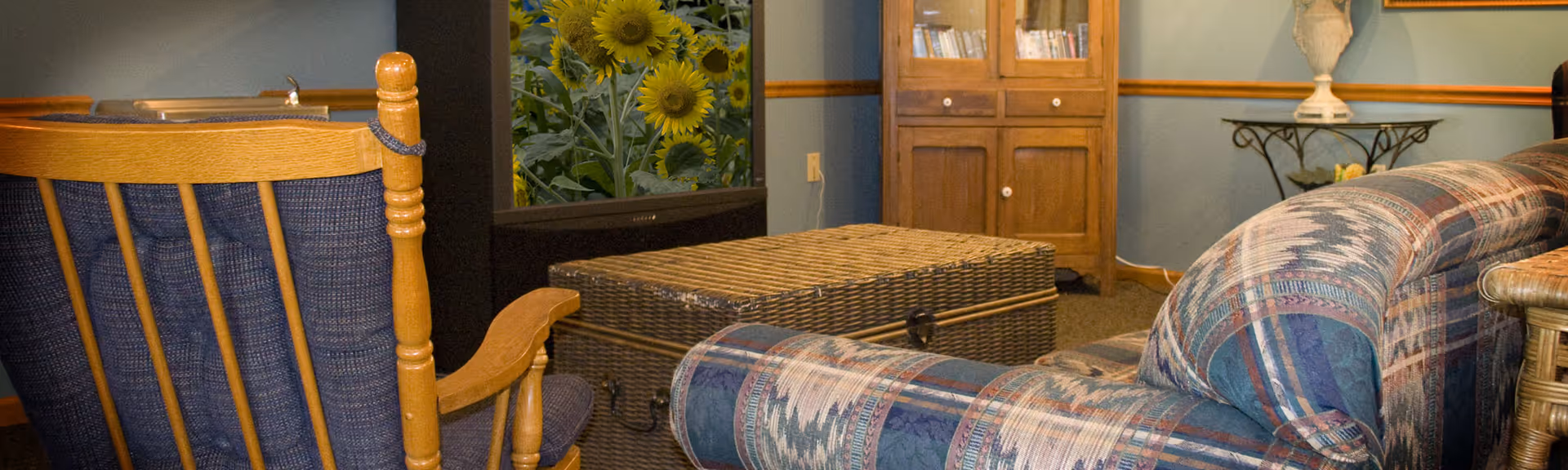 Cozy living room area with a patterned upholstered couch, a wooden rocking chair with blue cushions, a wicker chest used as a coffee table, a TV displaying an image of sunflowers, a wooden cabinet with books, and a decorative table with a lamp against a blue wall.