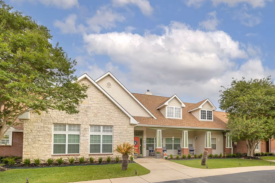 Exterior view of a single-story senior living facility building with beige stone and siding, a brown shingled roof, multiple windows, a covered porch with columns, and a well-maintained lawn with trees and shrubs under a partly cloudy sky.