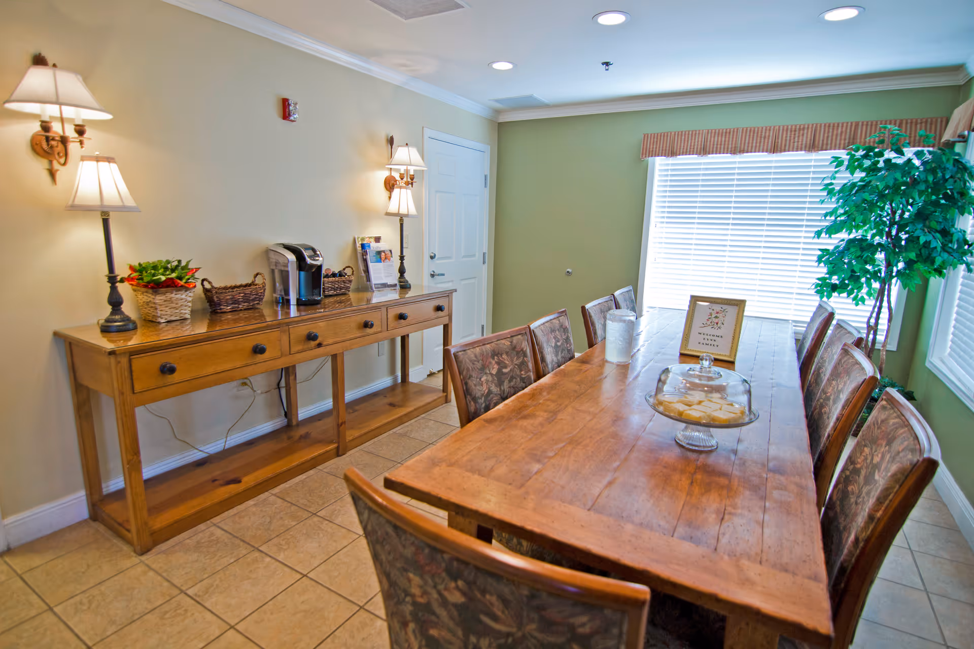 A dining room with a long wooden table surrounded by eight chairs with floral upholstery. On the table, there is a glass dome covering some food and a framed sign. To the left, a wooden sideboard holds a coffee maker, baskets, a plant, and two table lamps. The room has tiled flooring, light green walls, and a large window with blinds and a valance. A potted plant is in the corner near the window.