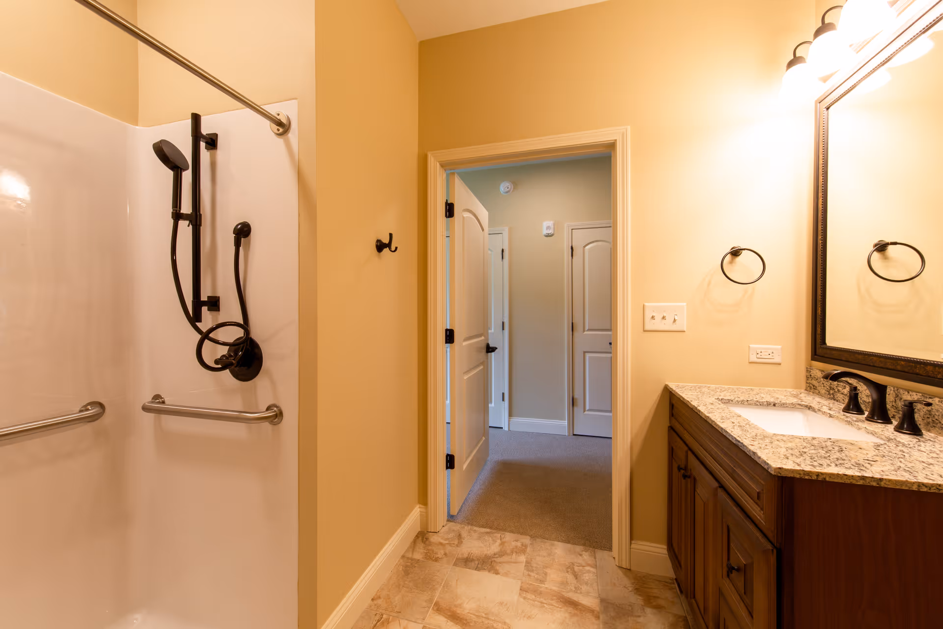 A bathroom with a walk-in shower featuring a handheld showerhead and grab bars, a wooden vanity with a granite countertop and sink, a large mirror with lighting above, and an open door leading to a carpeted hallway with two closed doors.