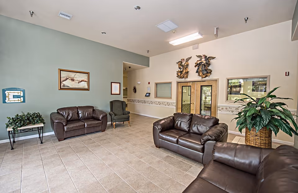 Spacious seating area with brown leather sofas, an armchair, potted plants, and decorative wall art in a tiled lobby.