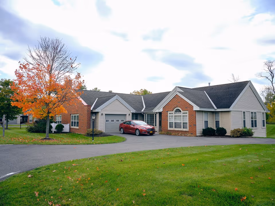 Single-story residential building with a combination of brick and siding exterior, a garage with a closed door, a red car parked in the driveway, and a tree with autumn-colored leaves on a green lawn under a cloudy sky.