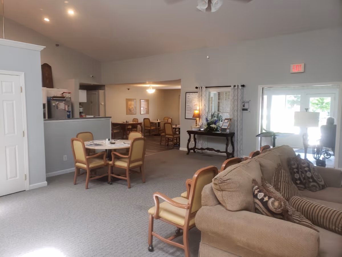 Interior view of a senior living facility showing a common area with beige sofas and patterned cushions in the foreground. There are several wooden chairs with yellow cushions around small round tables. In the background, there is a dining area with more tables and chairs, a kitchen area with white cabinets, and a side table with a lamp and decorative items. A person in a wheelchair is visible near the glass entrance door.