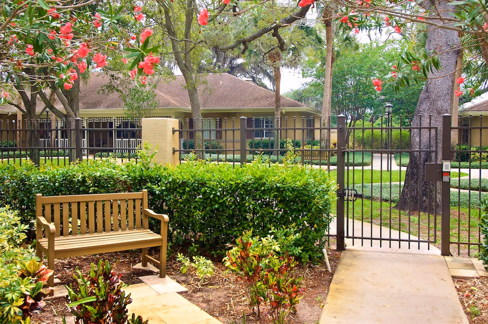 A peaceful outdoor garden area at Cottages at Belleair featuring a wooden bench surrounded by green bushes and flowering plants, with a metal gate and residential buildings in the background.