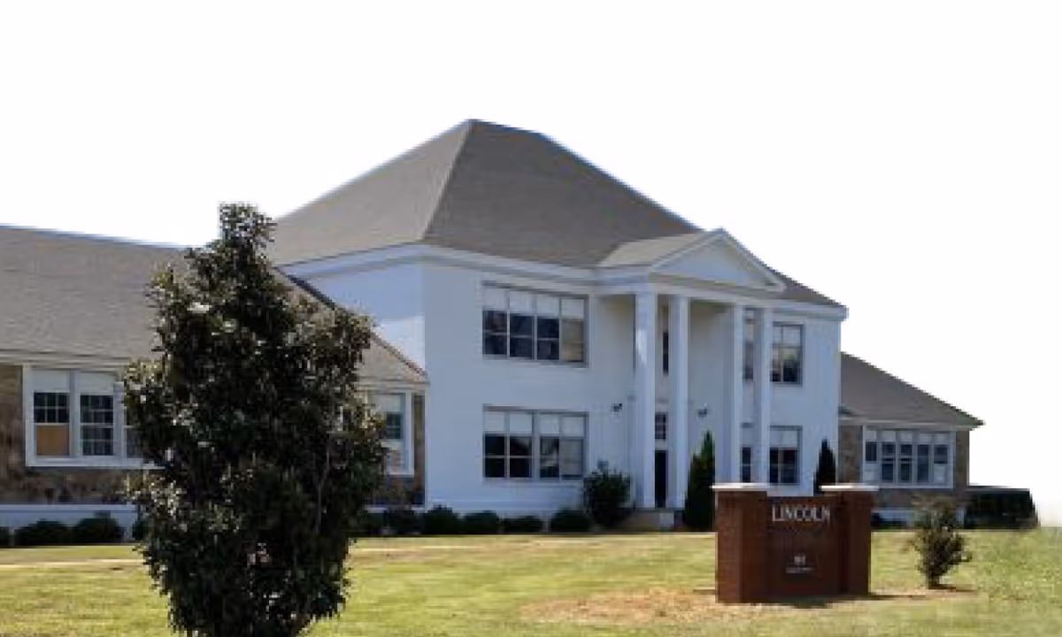 Exterior view of a large, two-story building with white walls and a gray roof, featuring tall white columns at the entrance. There is a green lawn and a tree in front of the building, along with a brick sign that reads 'LINCOLN'.