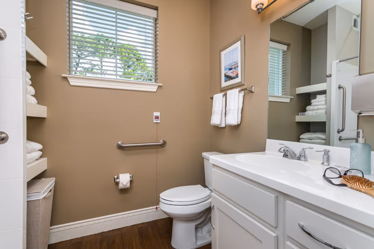 A clean and well-lit bathroom with beige walls and a window with white blinds. The bathroom features a white toilet, a white vanity with a sink, a large mirror, and a silver faucet. There are two white towels hanging on a towel rack above the toilet, a framed picture on the wall, and shelves stocked with neatly folded white towels. A grab bar is mounted on the wall next to the toilet, and a toilet paper holder is also visible. On the vanity countertop, there is a hairbrush, a pair of glasses, and a soap dispenser.