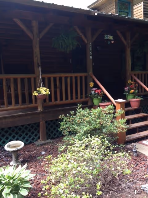 Front porch of a wooden cabin-style building with potted flowers on the railing and steps, surrounded by green bushes and garden plants. The porch has wooden railings and a hanging plant, with the number 3650 visible on a post.