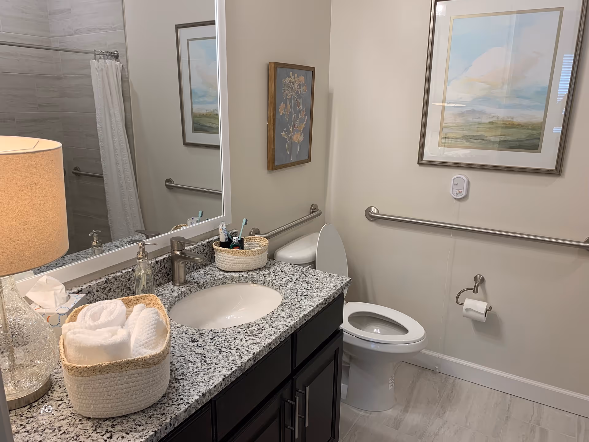 A clean and well-lit bathroom featuring a granite countertop with a sink, a lamp, a basket with rolled white towels, and a soap dispenser. There is a large mirror above the sink, a toilet with the lid open, a toilet paper holder, and grab bars on the walls. The bathroom has light-colored tiled flooring and two framed pictures on the walls.