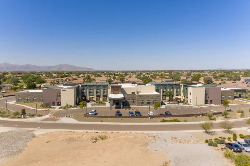 Aerial view of Surprise Health & Rehabilitation Center, a modern two-story building with a parking lot in front, surrounded by a suburban neighborhood with many houses and distant mountains under a clear blue sky.