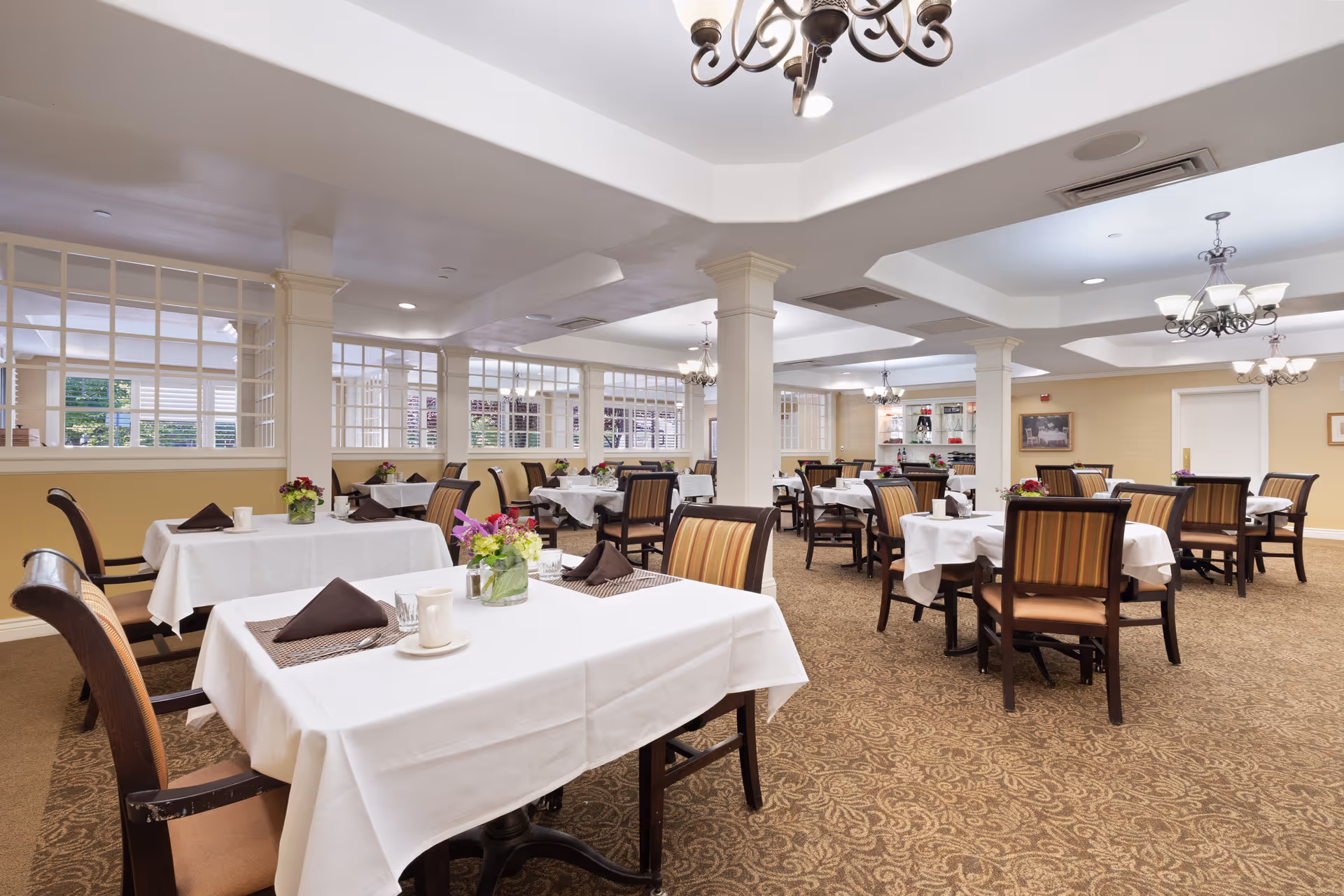 A spacious dining room in a senior living facility with multiple tables covered in white tablecloths, each set with brown napkins, cups, and small flower arrangements. The room features beige walls, patterned carpet, decorative chandeliers, and large windows with white frames allowing natural light to enter.