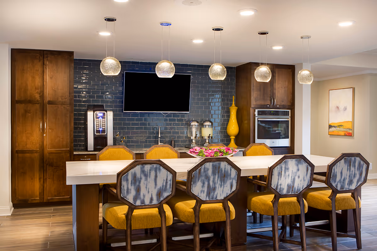 Modern kitchen area with a long white island countertop surrounded by six chairs with yellow cushions and patterned backs. The back wall features dark blue subway tiles, wooden cabinets, a built-in oven, a water dispenser, two beverage dispensers, a large yellow decorative vase, and a mounted flat-screen TV. Pendant lights hang from the ceiling above the island.