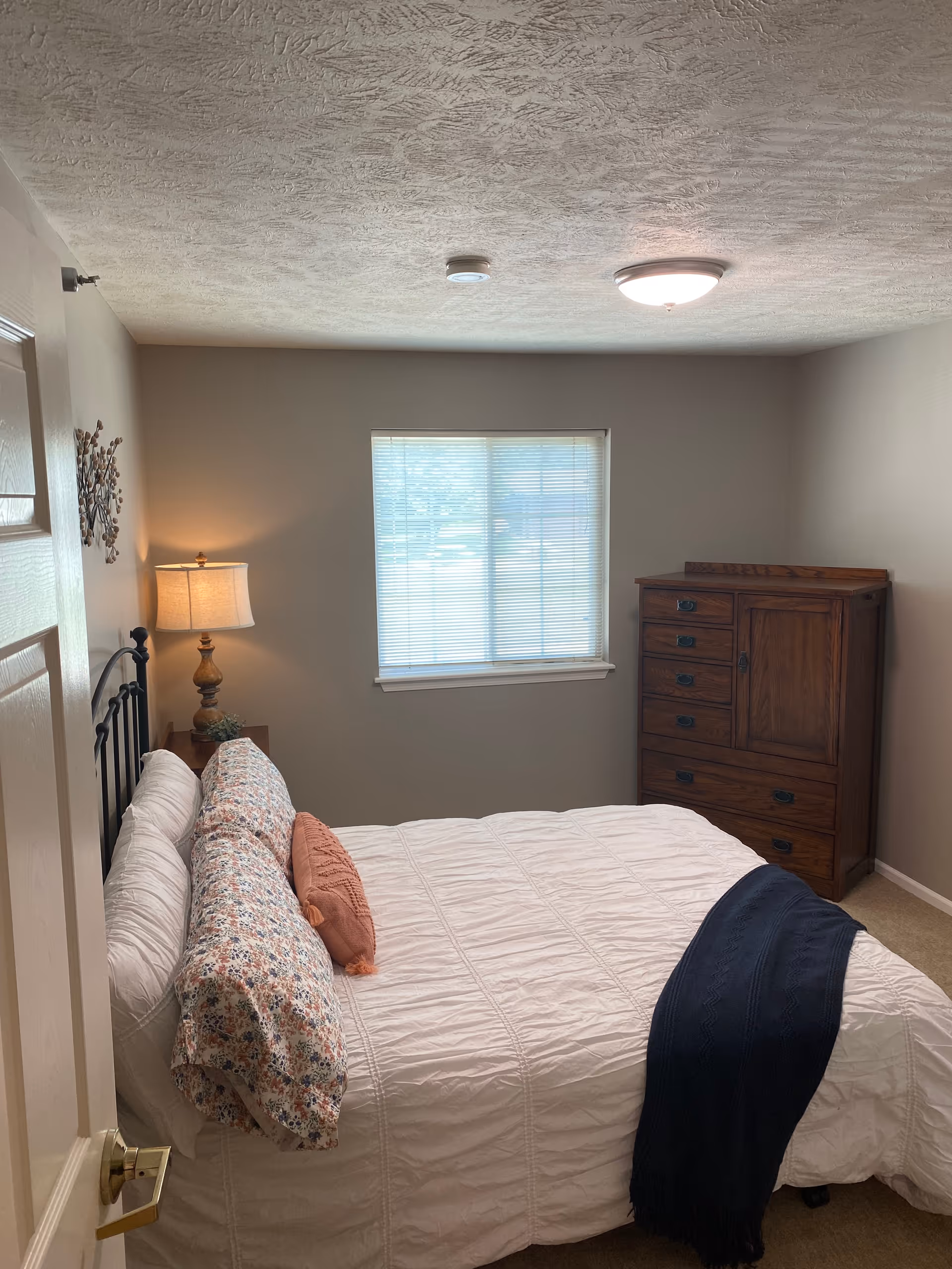 A cozy bedroom with a neatly made bed featuring white bedding, floral pillows, and a coral accent pillow. A dark blue throw blanket is draped across the foot of the bed. To the left of the bed is a wooden nightstand with a decorative lamp and a small plant. A window with closed blinds is centered on the far wall, and a wooden dresser with multiple drawers is positioned against the right wall. The walls are painted a neutral beige color, and the ceiling has a textured finish with a ceiling light fixture.