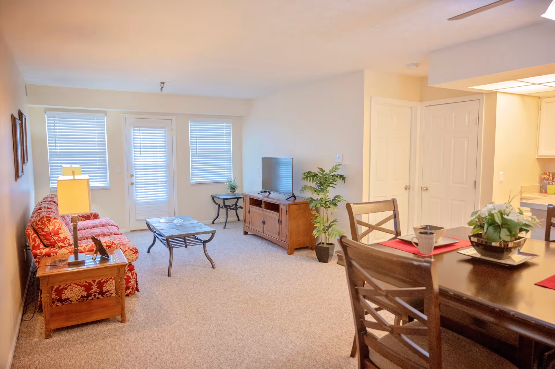 A bright living room with beige walls and carpet, featuring a red patterned sofa, a wooden coffee table, a wooden TV stand with a flat-screen TV, and two windows with blinds flanking a glass door. To the right, there is a dining area with a dark wooden table set with placemats, dishes, and a potted plant. The kitchen is partially visible in the background.