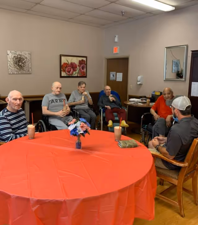 A group of elderly men sitting around a table covered with a red tablecloth in a room. Some men are in wheelchairs, and they are drinking beverages with straws. A vase with flowers is on the table. The room has beige walls with two framed pictures and a mirror. A man wearing a cap and mask is seated on a wooden chair facing the group.