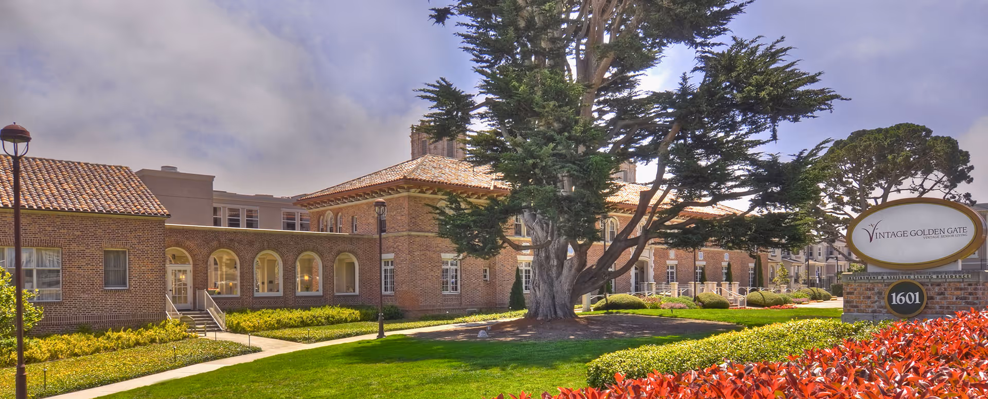 Exterior view of a senior living facility with brick buildings, a large tree in the center, well-maintained green lawns, shrubs, and a sign that reads Vintage Golden Gate Retirement & Assisted Living Residence at 1601.