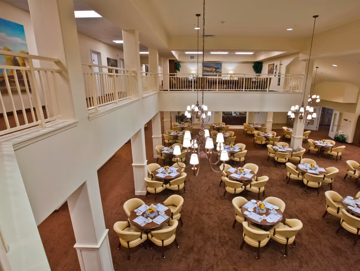 Two-story dining room with round tables set for meals, beige chairs and multiple hanging chandeliers.