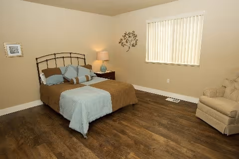 Furnished bedroom with a bed, nightstand and lamp, armchair, and a window with vertical blinds over wood-look flooring.