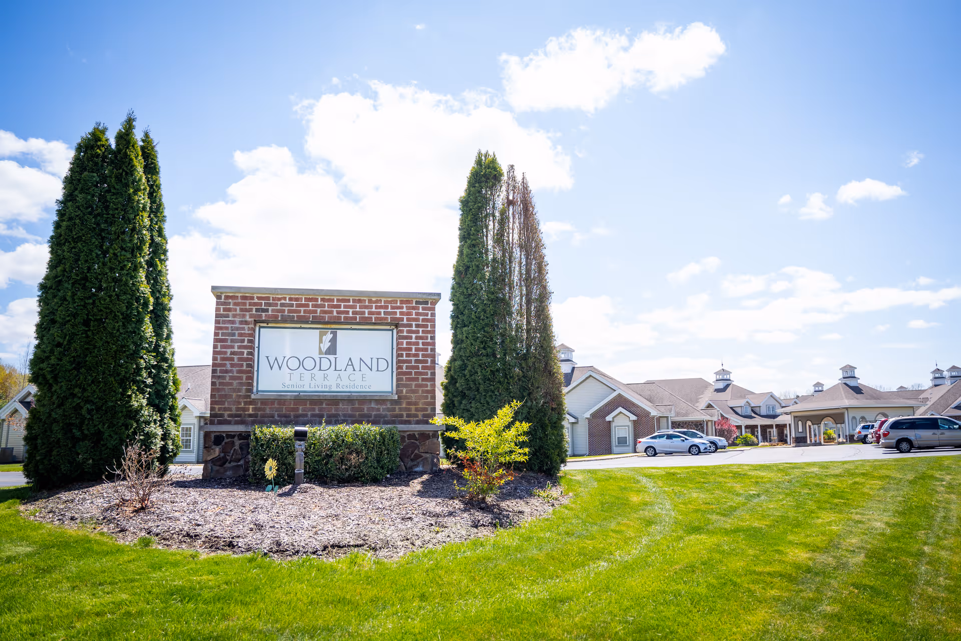 Front entrance sign for Woodland Terrace senior living residence on a landscaped lawn with the facility building and parked cars in the background.