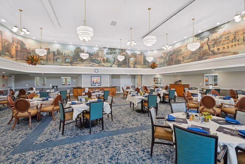 A spacious dining room with multiple round tables covered with white tablecloths, set with blue napkins, menus, and small flower arrangements. The room features a patterned carpet and a large mural depicting a scenic landscape with classical architecture along the upper walls. Several pendant lights hang from the ceiling, illuminating the area.
