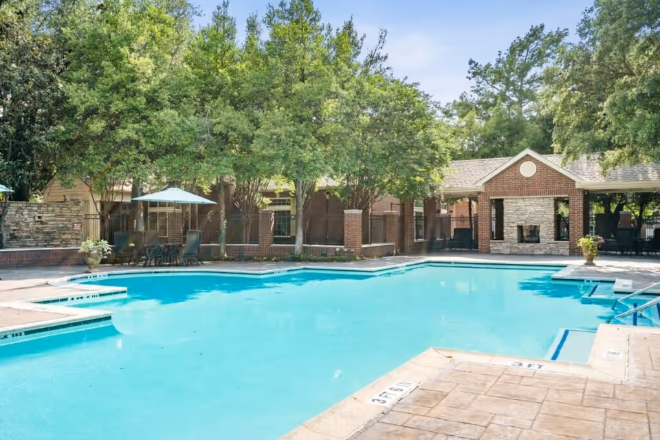 Outdoor swimming pool area with clear blue water surrounded by a concrete deck. There are several trees providing shade, patio tables with umbrellas, and chairs around the pool. In the background, there is a brick building with a covered seating area and a stone fireplace.