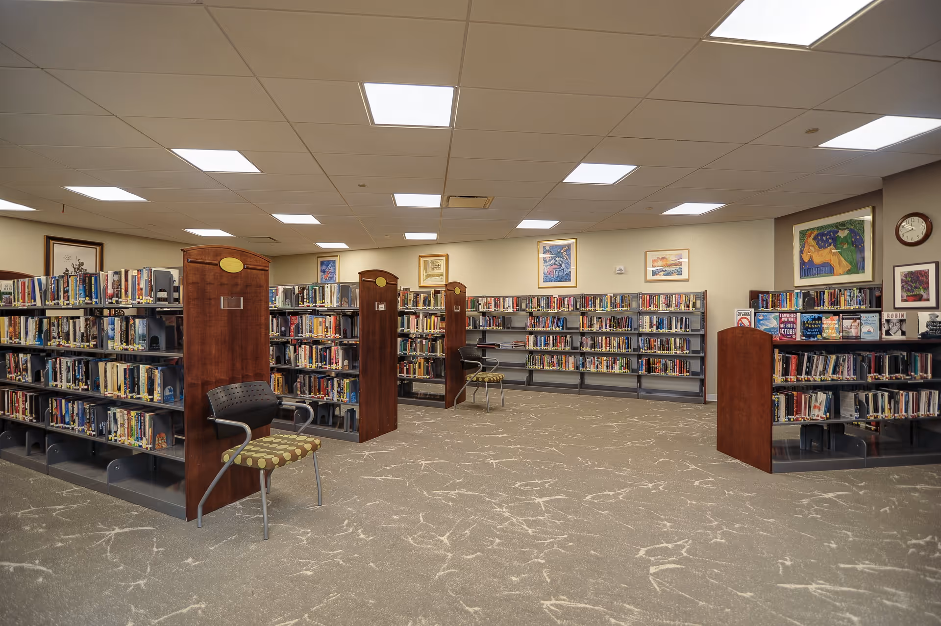 Interior view of a library room with multiple bookshelves filled with books. There are a few chairs placed near the shelves. The walls have framed artwork and a clock. The ceiling has recessed lighting panels.