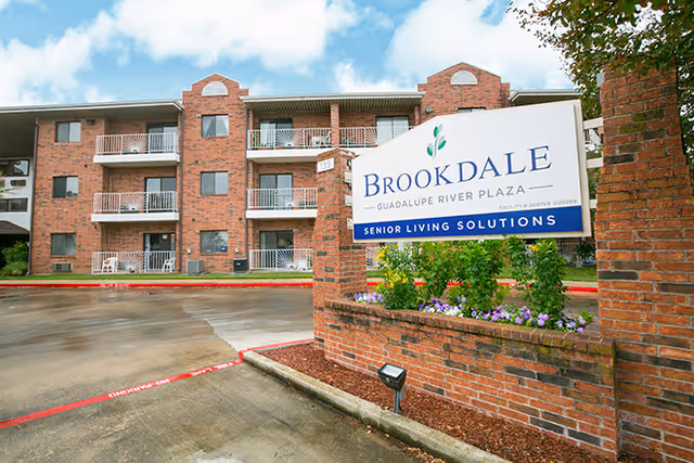 Exterior view of a three-story brick senior living facility with balconies. In the foreground, there is a brick sign that reads 'Brookdale Guadalupe River Plaza Senior Living Solutions' surrounded by flowers and greenery. The sky is partly cloudy.