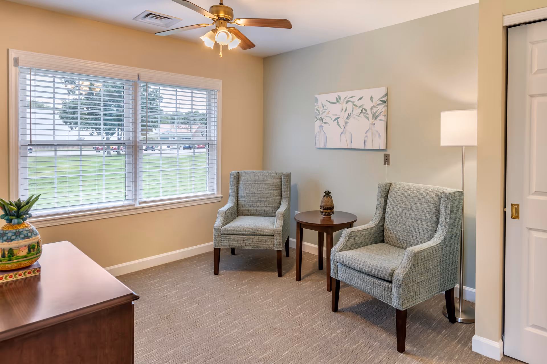 A cozy sitting area with two light blue upholstered armchairs positioned around a small round wooden table with a decorative pineapple on it. A floor lamp stands beside one chair, and a painting of three vases with leaves hangs on the wall. A window with white blinds lets in natural light, and a ceiling fan with lights is mounted above. A wooden cabinet with a colorful pineapple decoration is partially visible in the foreground.