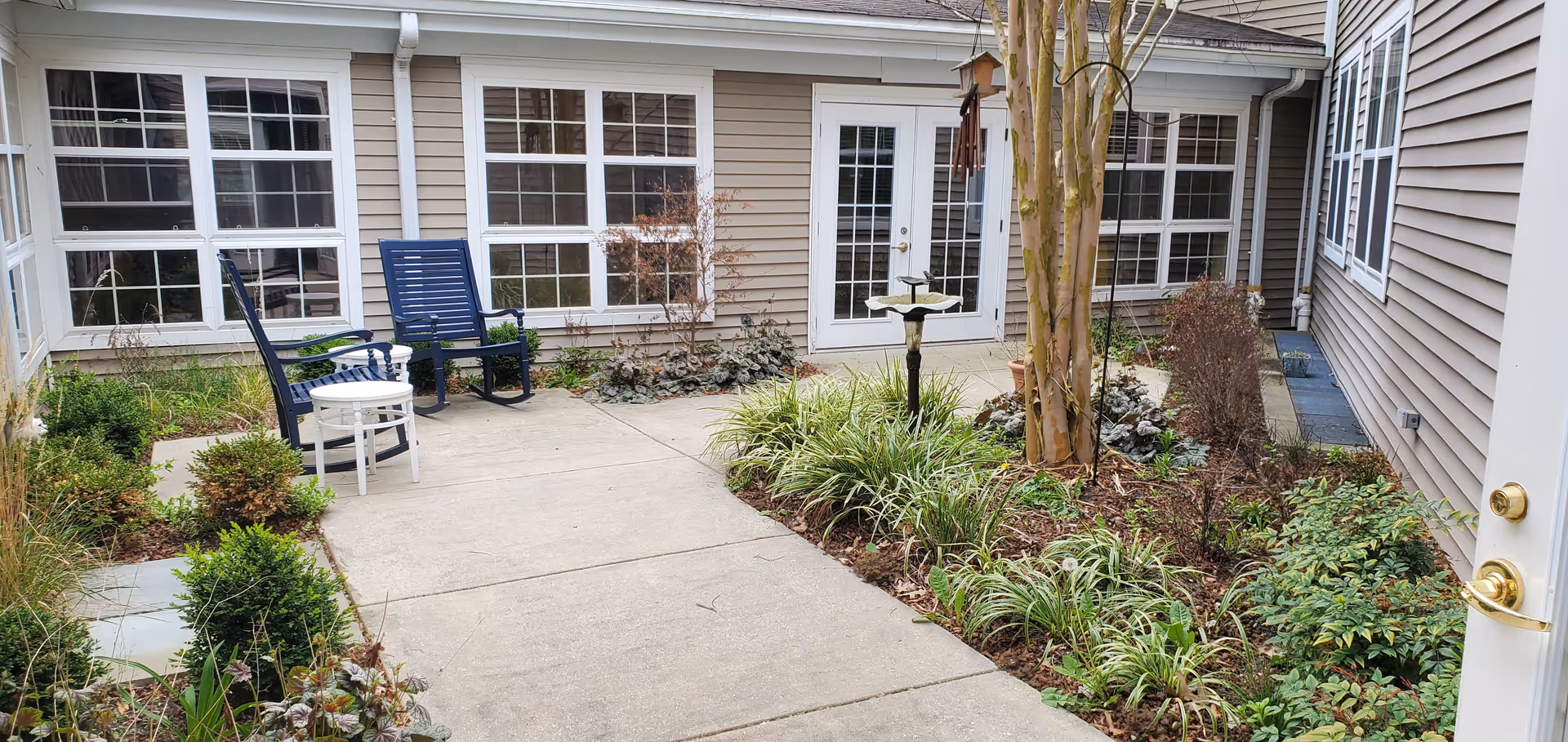 A small outdoor courtyard area with a concrete pathway surrounded by garden beds containing various plants and shrubs. Two blue rocking chairs and a small white table are placed on the left side near windows of the building. The building exterior is beige with white trim and multiple windows. A white door with glass panes is visible in the center background.