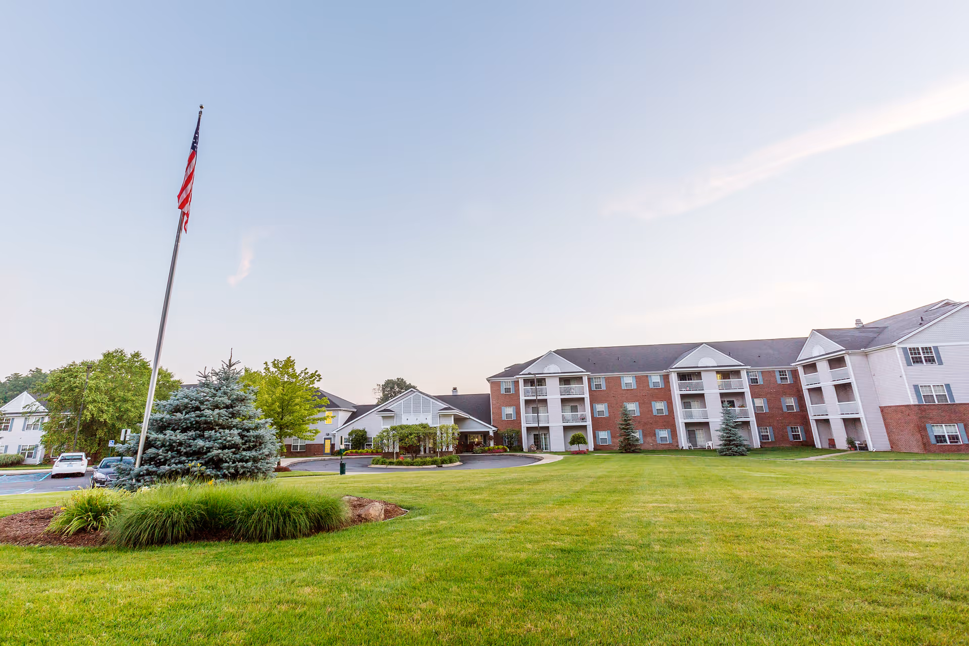 Exterior view of Independence Village of White Lake, showing a large green lawn with a flagpole flying the American flag, and a multi-story brick and white building in the background under a clear sky.