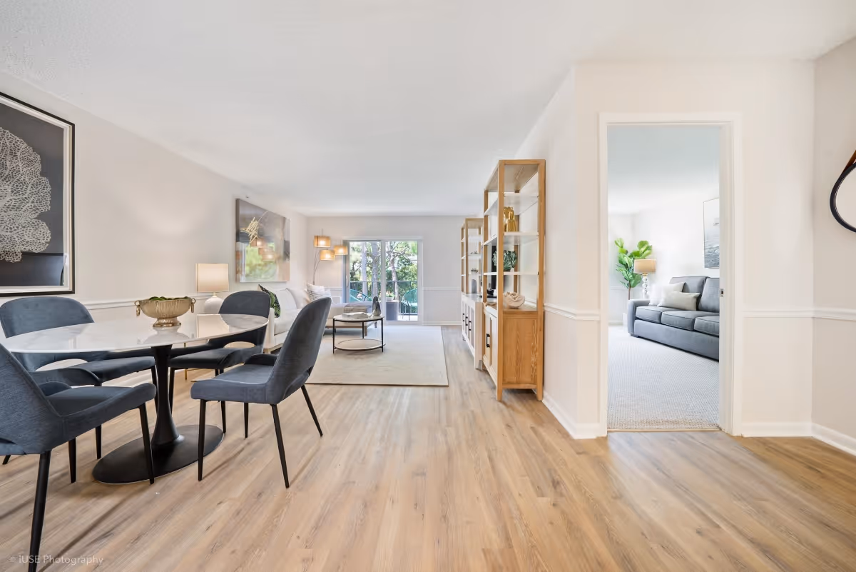 Bright and spacious interior of a senior living facility showing a dining area with a round glass table and four blue upholstered chairs on the left, a living room with a beige rug, coffee table, and sofa in the background, and a separate room with a gray couch and green plant visible through an open doorway on the right. The flooring is light wood, and the walls are painted white with decorative molding. Large windows at the back let in natural light and show greenery outside.