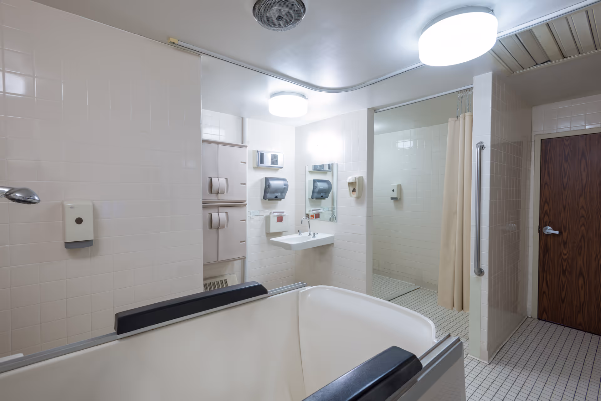 A clean, well-lit bathroom in a healthcare facility featuring a large bathtub with padded armrests, a wall-mounted sink with a mirror above it, a shower area with a curtain, tiled walls and floor, and a wooden door.