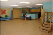 Open rehabilitation/therapy room with treatment tables, exercise balls, cabinets, and a practice staircase.