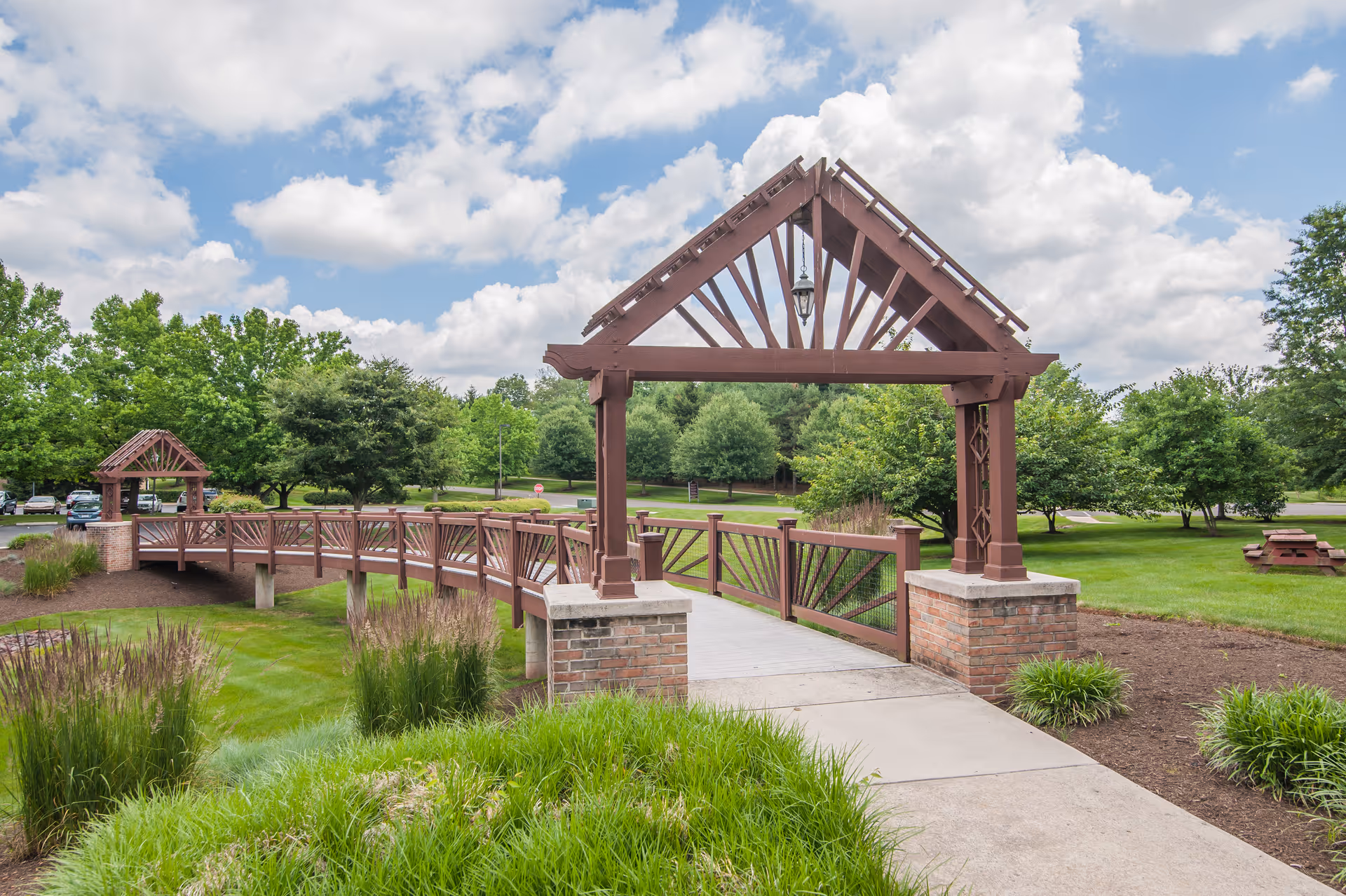 A wooden pedestrian bridge with decorative beams and brick pillars crosses over a small grassy area in a landscaped outdoor setting with trees, bushes, and a partly cloudy sky.