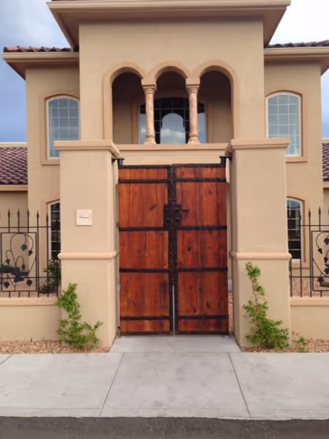 Front exterior view of a building with a large wooden gate set between beige stucco walls. The building has arched windows and a tiled roof, with some small plants growing near the base of the walls.
