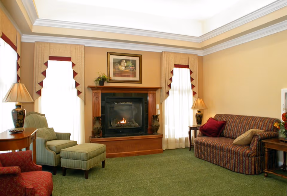 A cozy living room with a lit fireplace in the center, flanked by two tall windows with beige curtains featuring maroon accents. The room has a green carpet, a striped armchair with matching ottoman, a patterned sofa with red and beige pillows, two side tables with decorative lamps, and a framed painting above the fireplace.